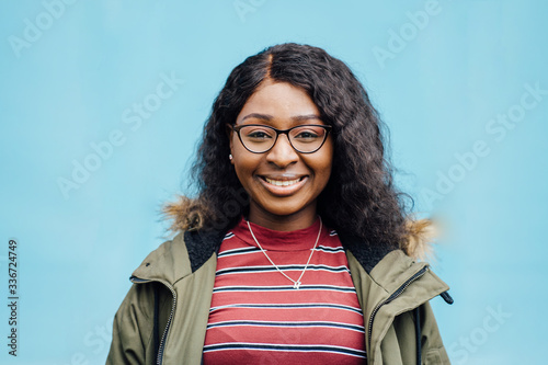 Portrait og young nigerian woman student wearing in khaki coat in eyeglasses on blue photo