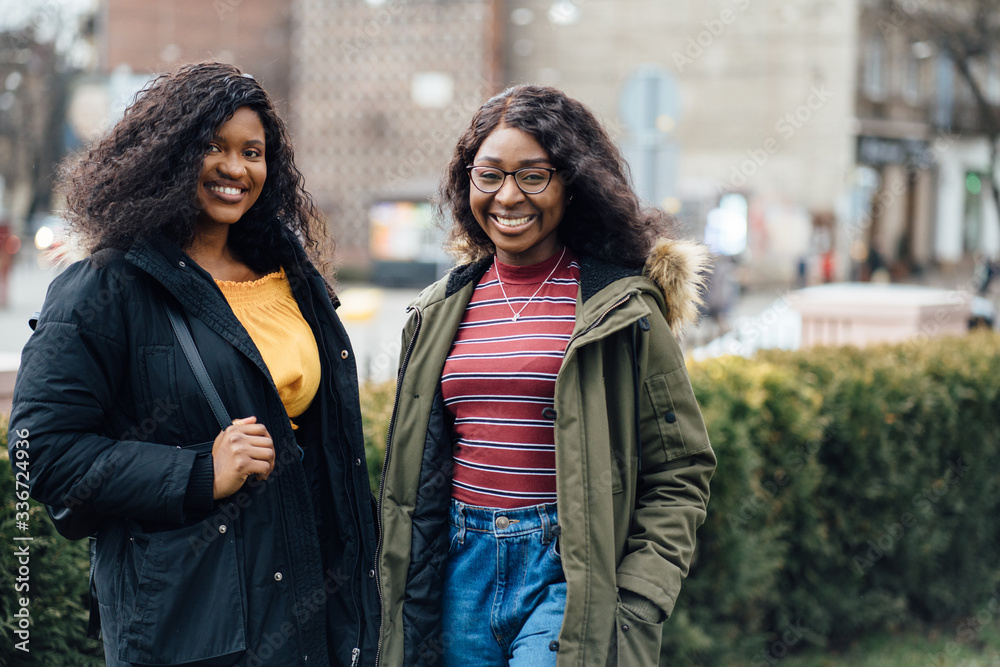 Portrait of two beautiful girls in urban backgrund, black and mixed women. Emotional dark skinned nigerian friends talking in city street, outdoor in spring cold weather.