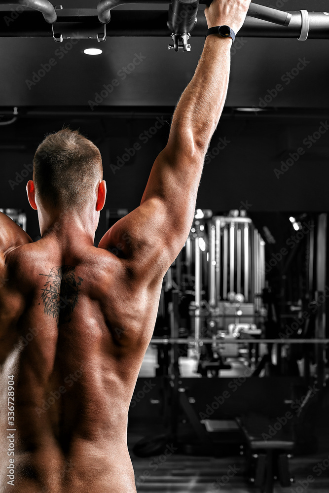 The athlete does pull-ups - chin in the gym, model with a sports body topless. Shot from the back, low key, close-up, copies of the space.