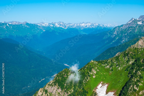 Mountain range with rocks and trees against a clear blue sky. Non-melted snow in the summer on a mountain range. Snow-capped mountain peaks on the horizon