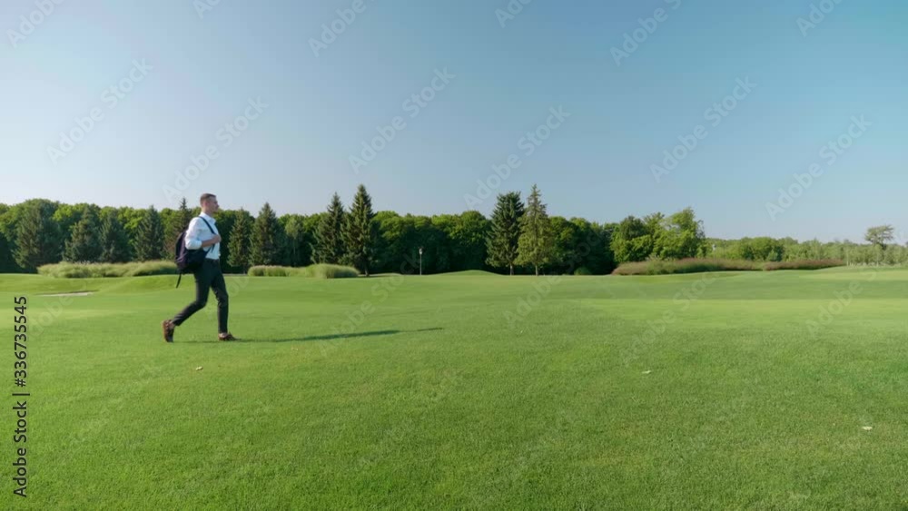 A young businessman with a backpack on his shoulder walks in the park. A man in a classic white shirt and trousers walks on green grass. 4K footage A young businessman with a backpack on his shoulder walks in the park. A man in a classic white shirt and trousers walks on green grass. 4K footage