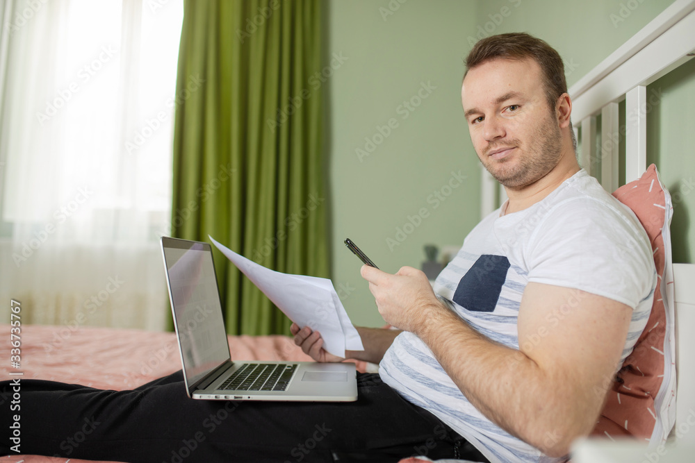 Adult man lying in his bed while he is working from home on his laptop looking on the camera. Papers or bills in his hand and a phone Adult man lying in his bed while he is working from home on his laptop looking on the camera. Papers or bills in his hand and a phone