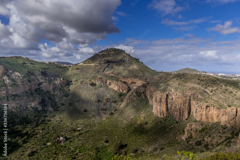 Gran Canaria nature around an erupting volcano rugged landscape full of hills and valleys during a sunny day full of clouds moving fast in the sky and shadows reflected on grassy soil.