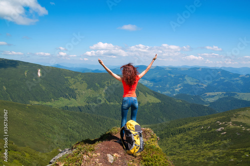 Happy young woman stands on a rock, her hands arms raised up to the blue sky
