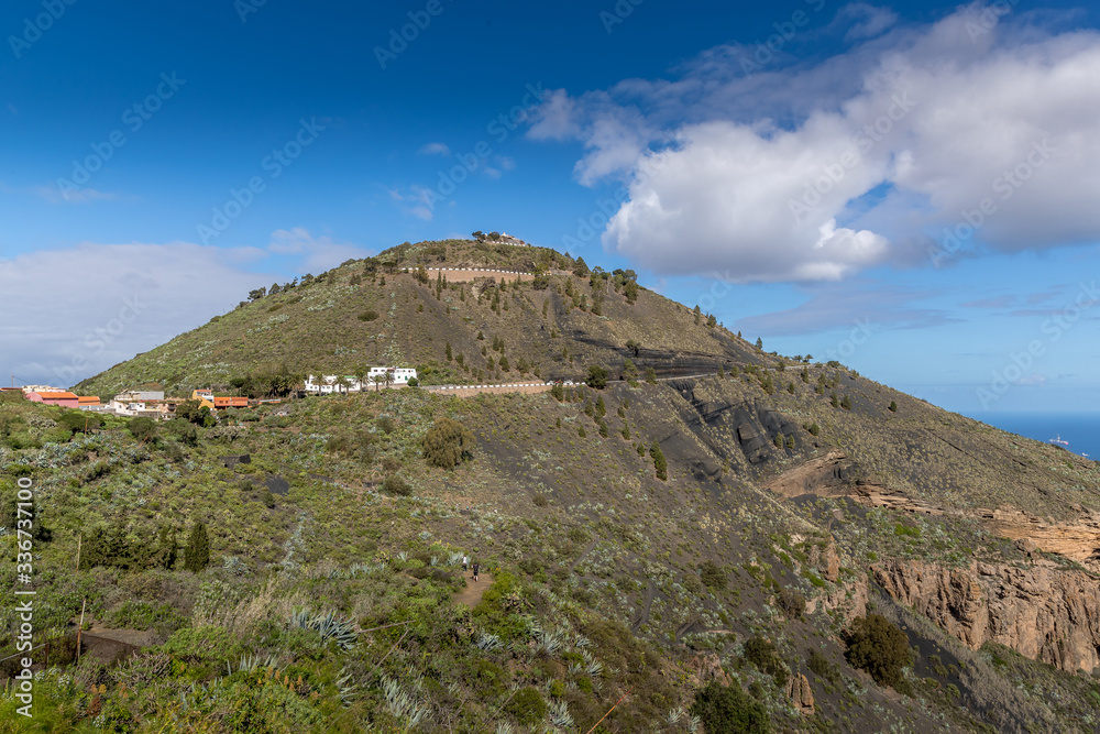 View of the hilltop on a mountainous landscape of volcanic origin along with lots of trees on the horizon, one can see the setting moon of the Gran Canary island