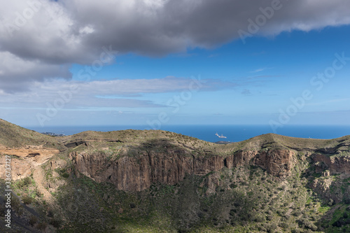 View of the hilltop on a mountainous landscape of volcanic origin along with lots of trees on the horizon, one can see the setting moon of the Gran Canary island