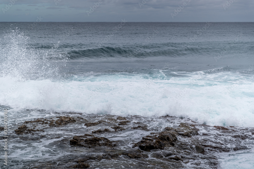 Fototapeta premium Waves crashing into a rocky shore full of large rocks and breaking water leaping to the sides with a pair of boats moored on the horizon at sea