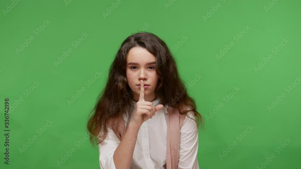 Be quiet, don't tell secret! Charming teenage brunette girl in white shirt putting finger on lips, showing silence gesture and looking mysteriously. indoor studio shot isolated on green background Be quiet, don't tell secret! Charming teenage brunette girl in white shirt putting finger on lips, showing silence gesture and looking mysteriously. indoor studio shot isolated on green background
