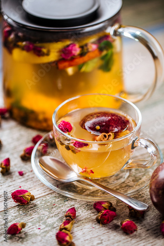 Closeup on the healthy herbal yellow apple tea with passion fruit and cinnamon in a glass teapot and mug on the wooden background decorated with roses, vertical
