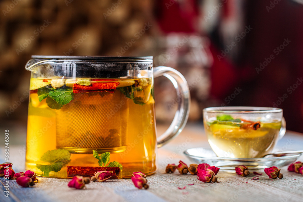 Closeup on the healthy herbal yellow apple tea with mint and cinnamon in a glass teapot and mug on the wooden background decorated with roses, horizontal Closeup on the healthy herbal yellow apple tea with mint and cinnamon in a glass teapot and mug on the wooden background decorated with roses, horizontal