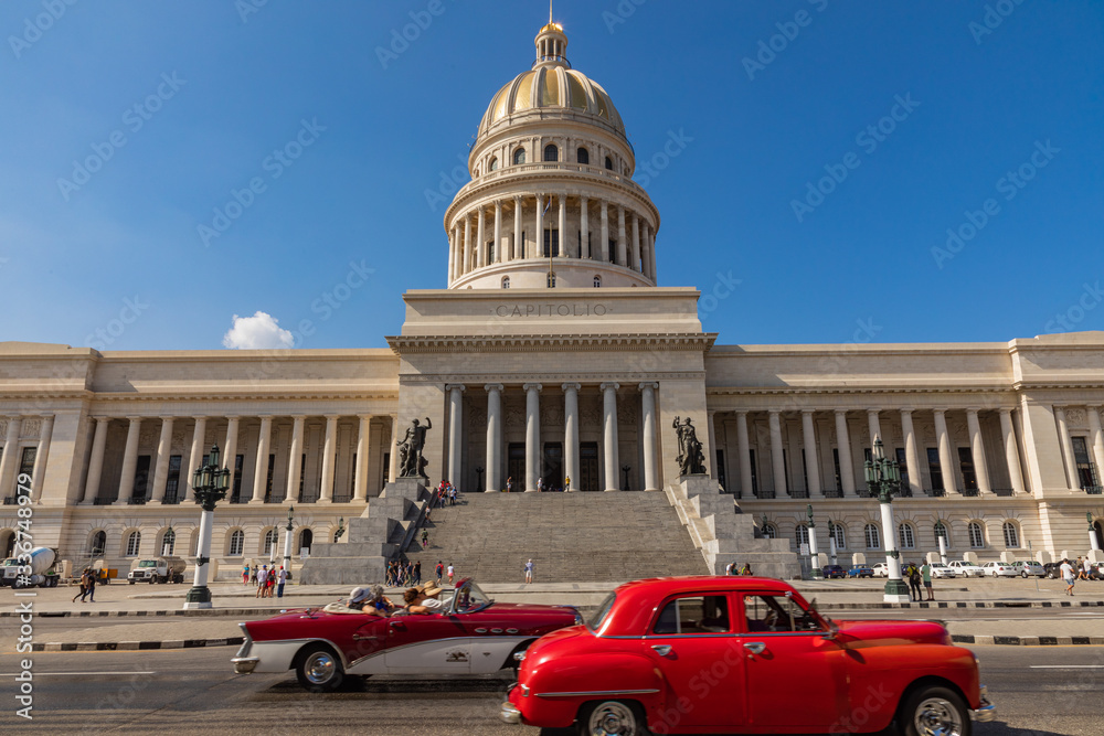 Brightly colored classic American cars serving as taxis pass on the main street in front of the Capitolio building in Central Havana, Cuba.