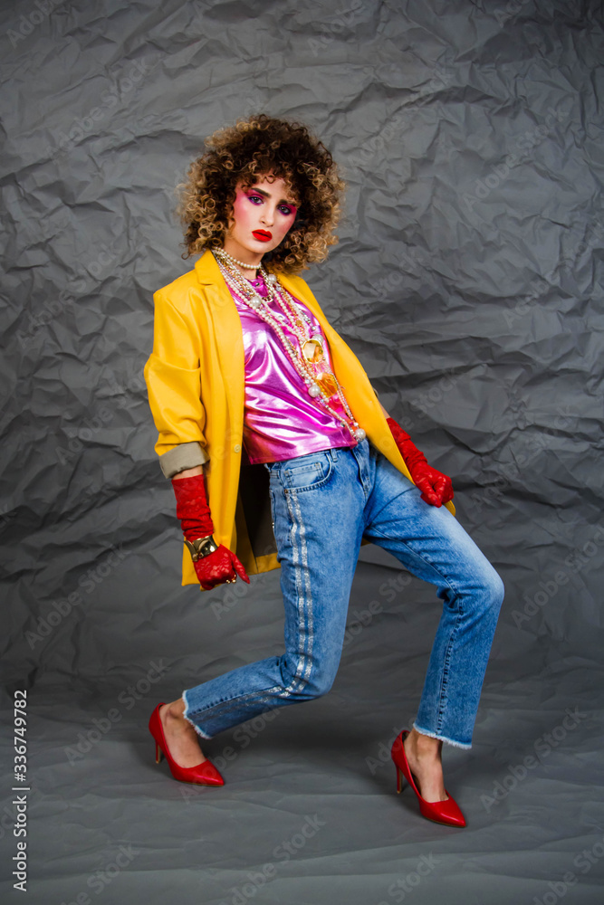 Girl in a yellow jacket and blue jeans with an afro hairstyle. Fashion of the eighties, disco era. Studio photo on gray background.