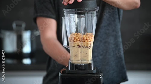 Young athlete man mixing hummus in a modern grey kitchen. Preparation of chickpea spread hommos in blender. Lentil dip, with spices and onion in liquidizer on grey kitchen background with pot. Full HD