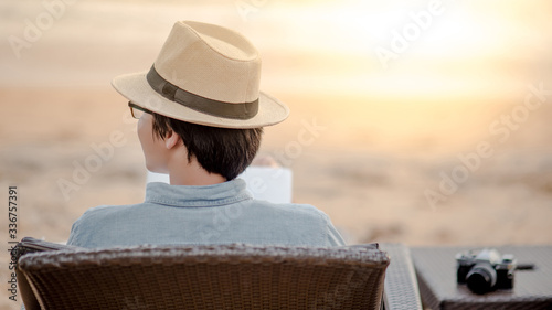 Asian man sitting on beach chair with his film camera at tropical island beach looking at the sea when sunset. Summer holiday or vacation travel concepts
