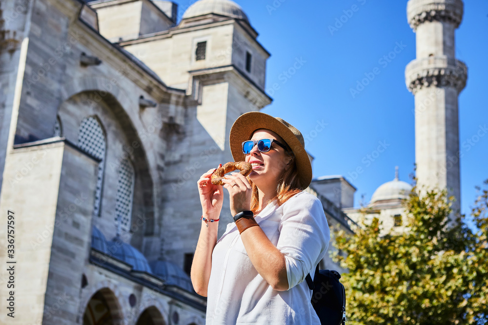 Happy attractive woman tourist in hat, eating Simitci (a round bagel with sesame seeds) against the background of Suleymaniye mosque, Istanbul,Turkey. Religion and travel concept.