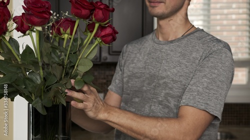 Young handsome man with a little stubble in a gray T-shirt straightens fixing and sniffs a beautiful bouquet of red roses that stands in a blue vase on a granite table in a fashionable kitchen