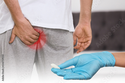 Doctor holding suppository for hemorrhoid treatment and man suffering from pain, closeup