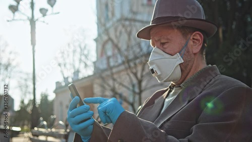 Pensioner elderly man using smartphone on bench in city street. Old grandfather with respiratory mask reading news staying outside. People on quarantine. photo