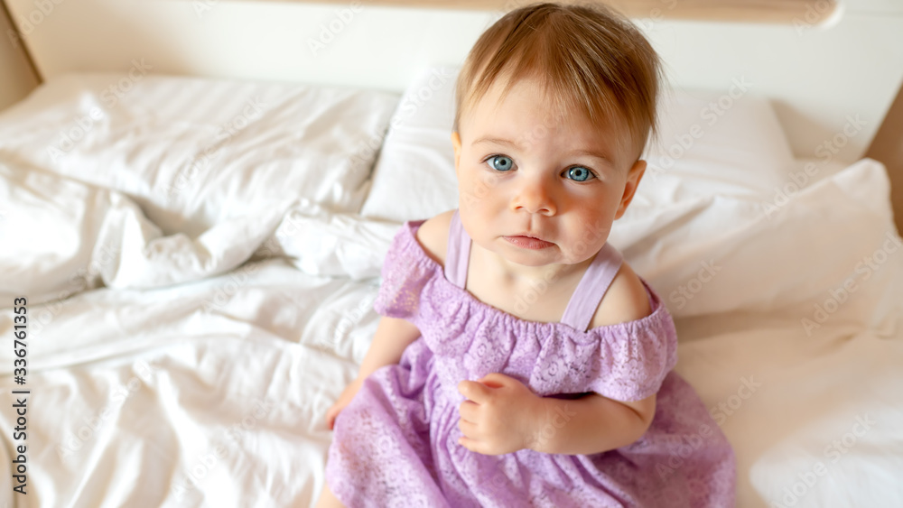 Close up portrait of sad blue-eyed toddler. Baby looks to the camera sitting on the bed. Girl in dress looking up. Close up portrait of sad blue-eyed toddler. Baby looks to the camera sitting on the bed. Girl in dress looking up.
