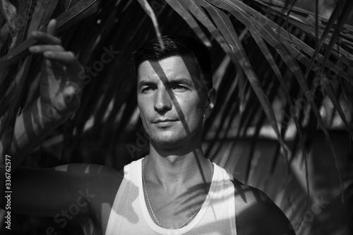 A young man with dark hair under a palm leaf with chiaroscuro from the branches on his face. A man in a tropical garden in India-a close-up portrait.