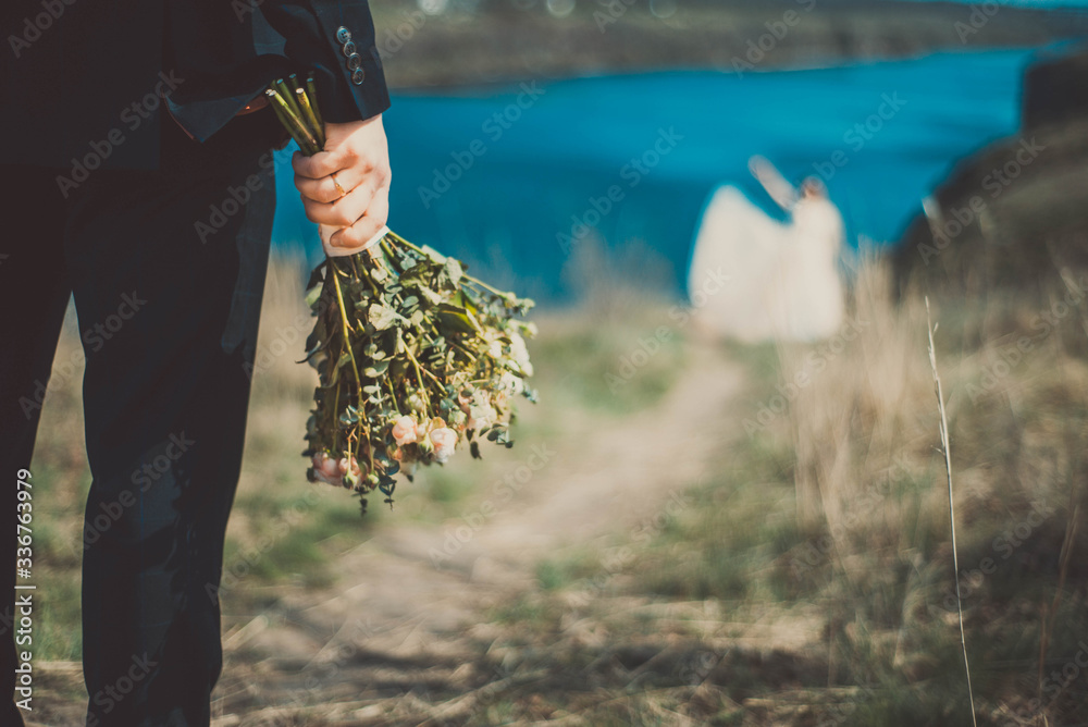 Obraz premium in the foreground in focus, the bride holding a bouquet of flowers, in defocus blur the bride hurries to the groom along the path. Against the background of the natural landscape.