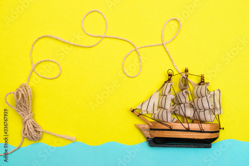 Fototapeta Naklejka Na Ścianę i Meble -  Toy ship is floating on the blue paper-cut waves, rope is twisted by loops above a ship on the yellow background.Concept summer and travel flat lay, copy space for mock-up