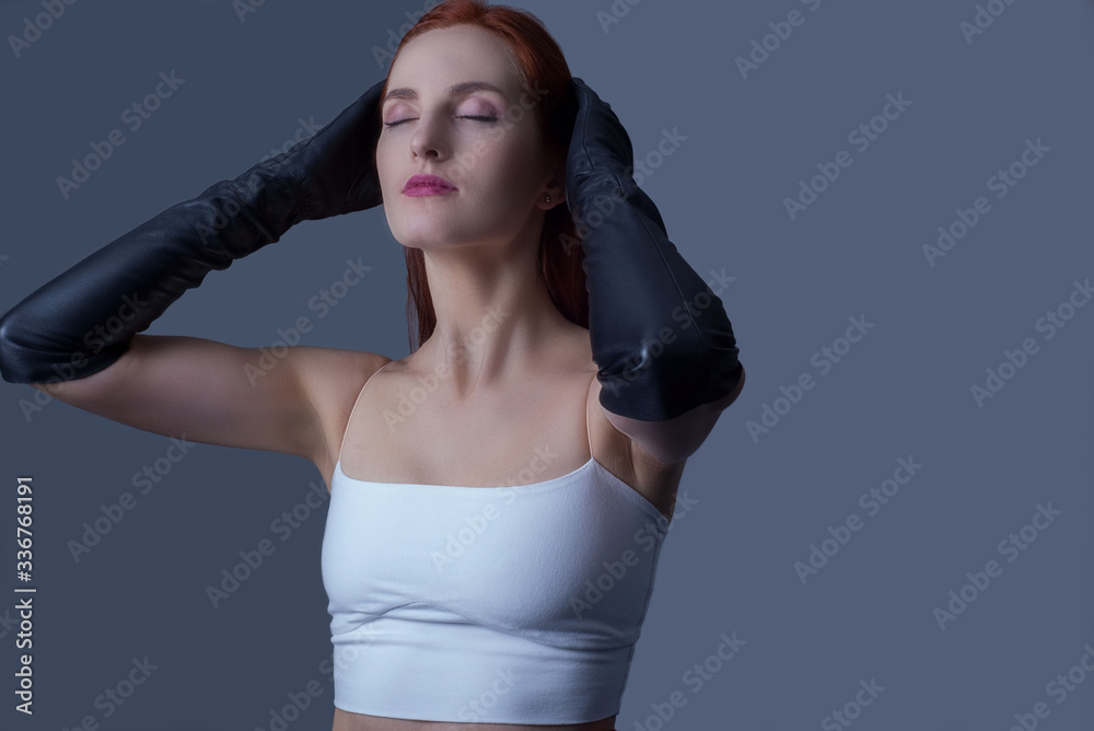 Naklejka premium Portrait of a young beautiful red-haired girl of thirty-five years old, in black leather gloves and a white tank top, touching her head with her hands, the model poses for a catalog magazine.