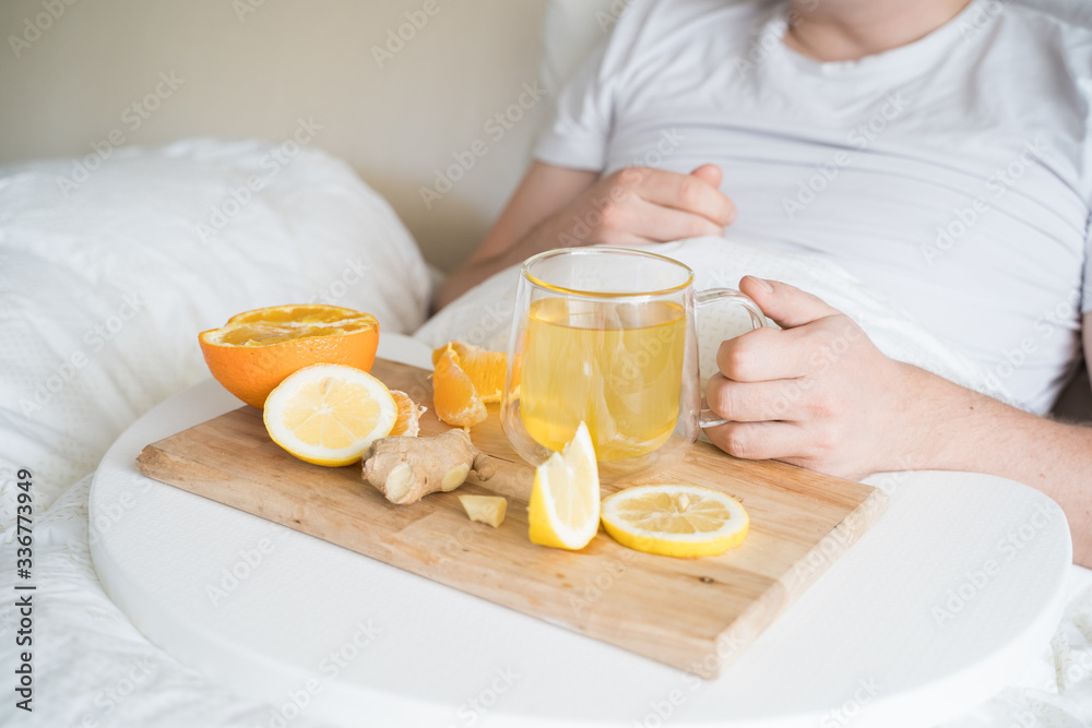 Cup with antipyretic drugs for colds,flu.Sick man in bed. Tea with citrus vitamin C,ginger root,lemon,orange.Wooden tray. Home self-treatment.Medical quarantine antiviral covid-19 coronavirus therapy Cup with antipyretic drugs for colds,flu.Sick man in bed. Tea with citrus vitamin C,ginger root,lemon,orange.Wooden tray. Home self-treatment.Medical quarantine antiviral covid-19 coronavirus therapy
