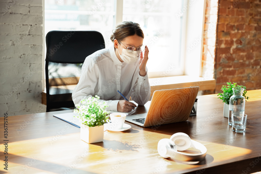 Meeting. Woman working in office alone during coronavirus or COVID-19 quarantine, wearing face mask. Young businesswoman, manager doing tasks with smartphone, laptop, tablet has online conference. Meeting. Woman working in office alone during coronavirus or COVID-19 quarantine, wearing face mask. Young businesswoman, manager doing tasks with smartphone, laptop, tablet has online conference.