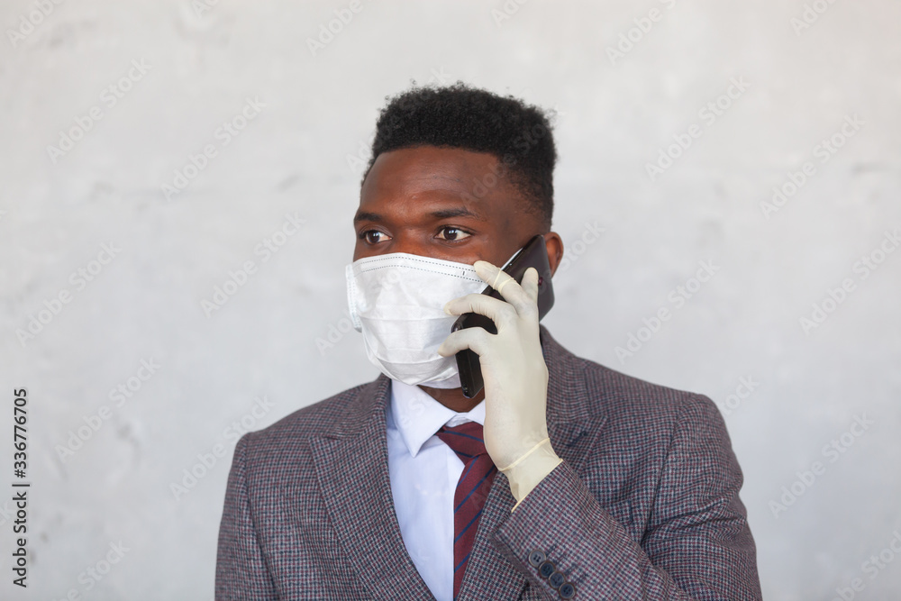 COVID-19. Handsome African black man in a suit is using medical mask and holding his mobile phone with a hand in gloves. Measures to prevent coronavirus infection and the development of a pandemic COVID-19. Handsome African black man in a suit is using medical mask and holding his mobile phone with a hand in gloves. Measures to prevent coronavirus infection and the development of a pandemic