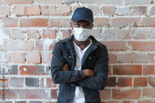 COVID, COVID-19, coronavirus, pandemic Young african black man wearing medical mask is standing in a cap with crossed arms shocked with coronavirus 2019 danger on background of ruined red brick wall