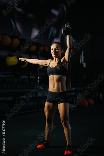 Fitness young woman with perfect muscular body in black sportswear is lifting kettlebell overhead during weight training workout. Concept of healthy lifestyle and workouts in a modern dark gym.
