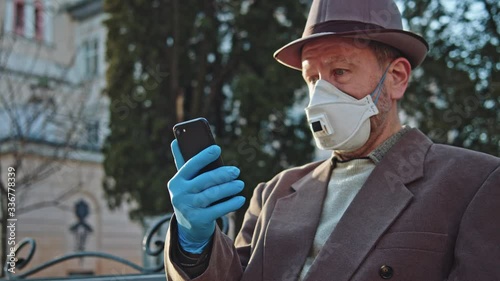 Old shocked man browsing content on smartphone reading news about coronavirus pandemic sitting on bench in the street. Pandemic, quarantine, COVID-19. photo