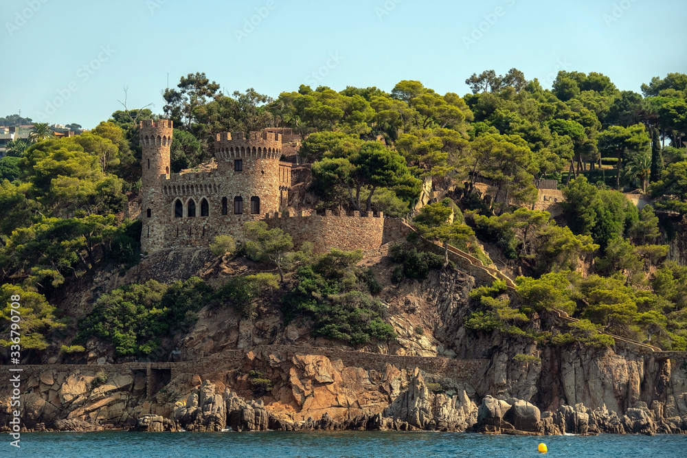 Ancient stone fortress with towers on a rocky cliff among green trees over the blue sea