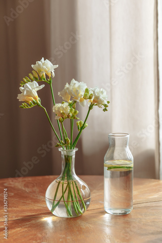 White flowers with a green stem in a transparent vase with water and a vessel with lemon on a table on a beige background.