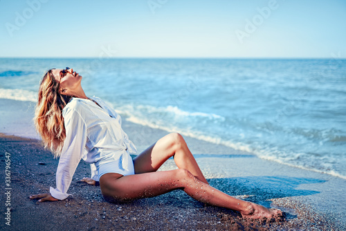 portrait of beautiful sexy caucasian sunbathed woman in sunglasses with long hair in swimsuitand white t-shirt lying on summer beach.