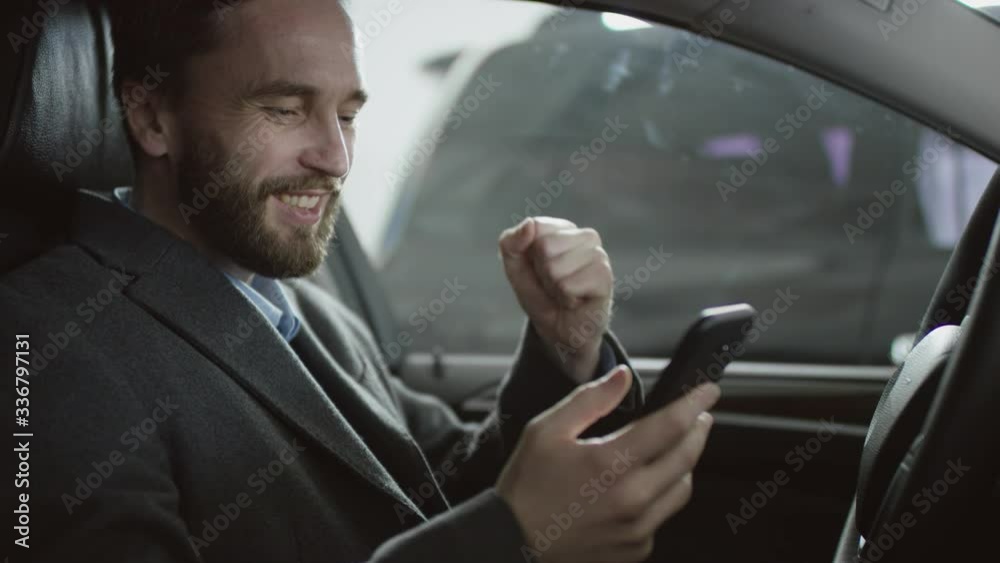 A businessman is surfing the internet sitting in the car on a driver's seat and using his phone. Businessman receiving good news while sitting in a car.