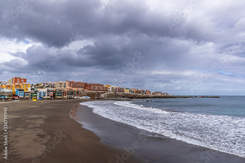 Black sand beach and with houses on rocks around coast with moving clouds and ocean during sunny day and a handful of people walking along the beach from Gran Canary Island. © Lukas