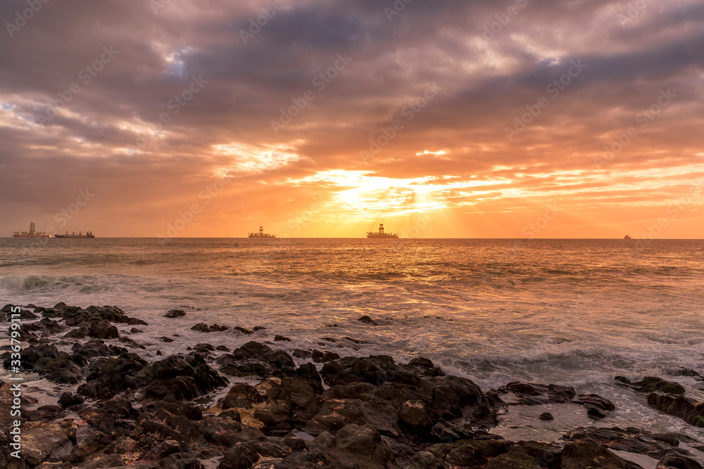Obraz premium Sunrise from a stony shore to boats moored on the horizon with the rising sun and pesky shining through the clouds to the ocean with moving clouds from the Grand Canary Island.