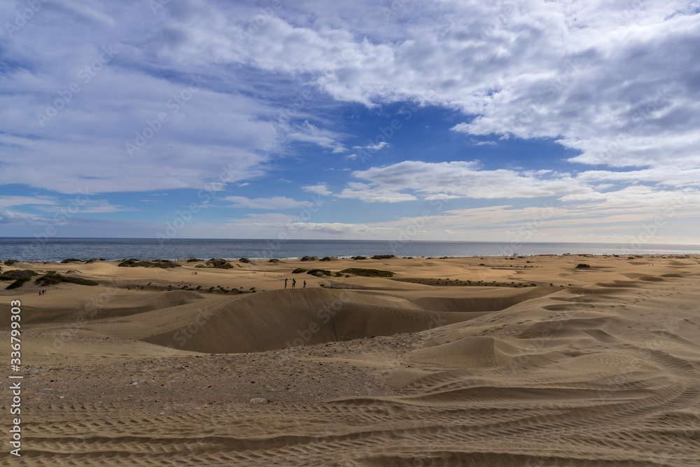 People passing through sand dunes overlooking the ocean on the horizon during a sunny day with thick clouds moving in the sky from the Grand Canary Island