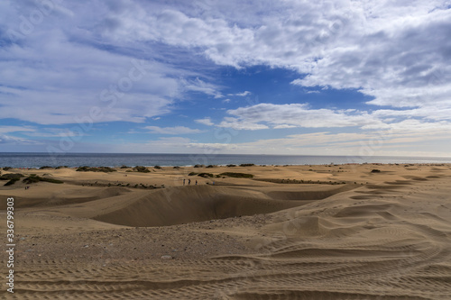 People passing through sand dunes overlooking the ocean on the horizon during a sunny day with thick clouds moving in the sky from the Grand Canary Island