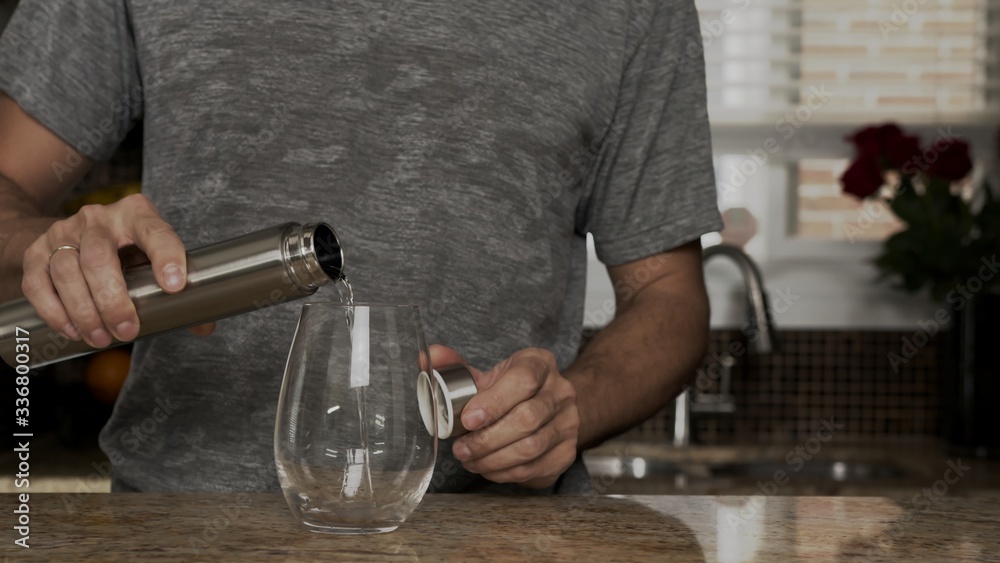 Young man in a gray T-shirt pours water into a glass from a stainless steel thermos or plastic bottle in a beautiful fashionable kitchen. healthy lifestyle. Handsome man is thirsty.