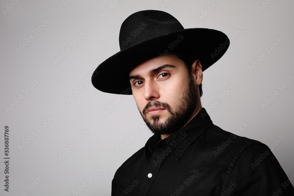 Side portrait of bearded hipster wearing black fedora hat and shirt looking at camera. Side portrait of bearded hipster wearing black fedora hat and shirt looking at camera.
