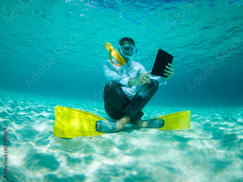 Snorkelling businessman in shirt and tie and matching fins using a tablet computer sitting underwater in tropical turquoise sea