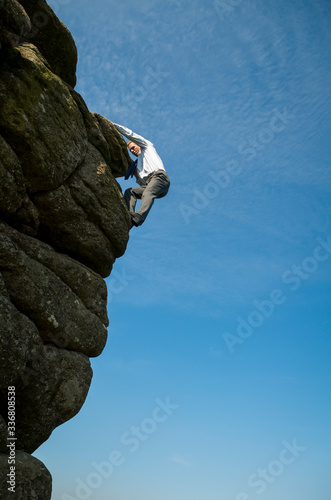 Brave businessman in shirt and tie climbing up the side of a dangerous rocky cliff outdoors in blue sky