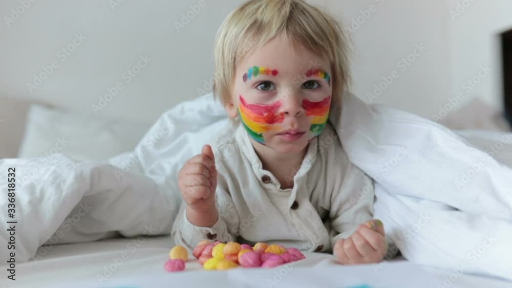 Beautiful blond toddler boy with rainbow painted on his face and messy hands, smiling happily Beautiful blond toddler boy with rainbow painted on his face and messy hands, smiling happily