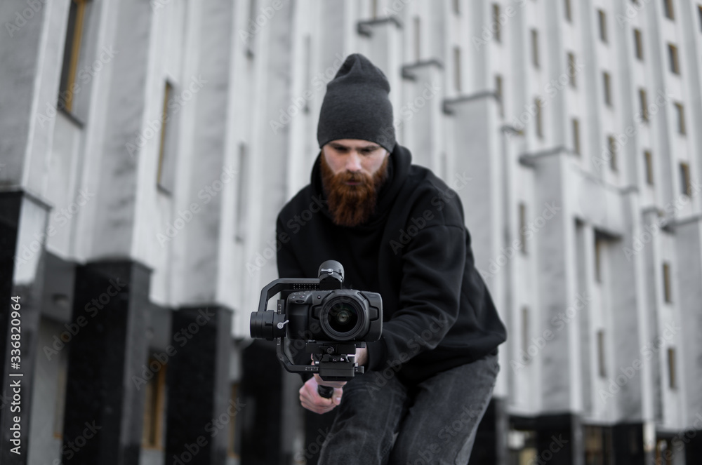 Bearded Professional videographer in black hoodie holding professional camera on 3-axis gimbal stabilizer. Filmmaker making a great video with a professional cinema camera. Cinematographer. Bearded Professional videographer in black hoodie holding professional camera on 3-axis gimbal stabilizer. Filmmaker making a great video with a professional cinema camera. Cinematographer.