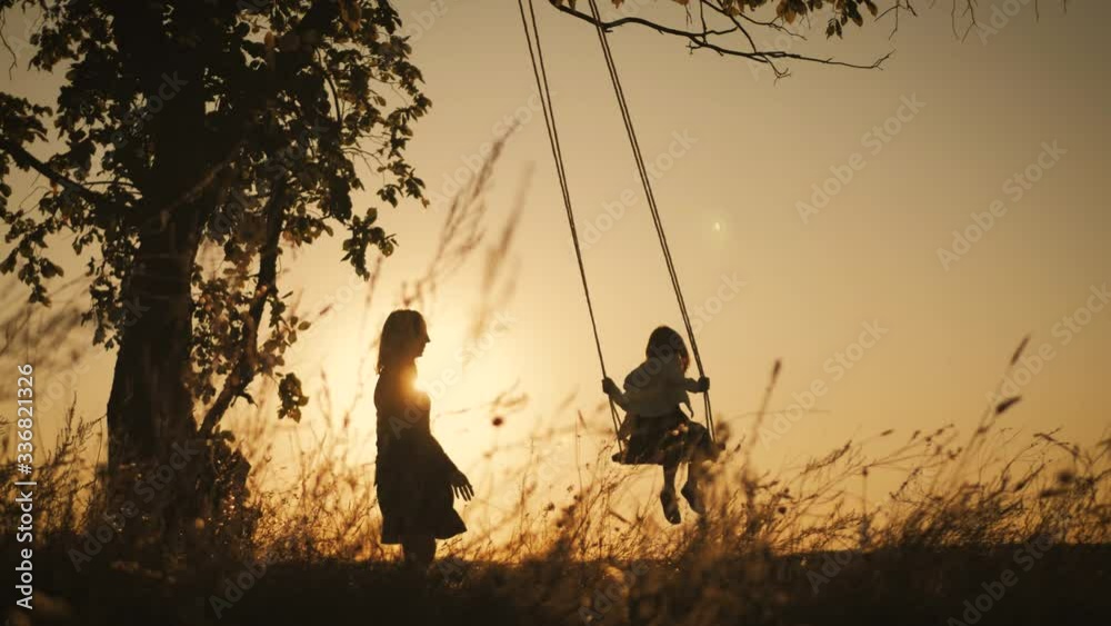 Silhouette of happy young mother and little daughter on a swing at sun light. Pretty girl sitting on a wooden swing and looking at golden sunset. Happy family concept. Stock ビデオ | Adobe Stock