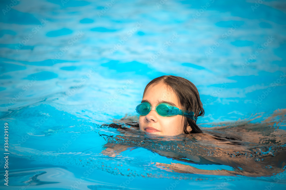 Charming girl teenager wearing waterproof glasses for the pool swims in the clear warm and blue water during the summer holidays. Concept of physical activity for children. Advertising space