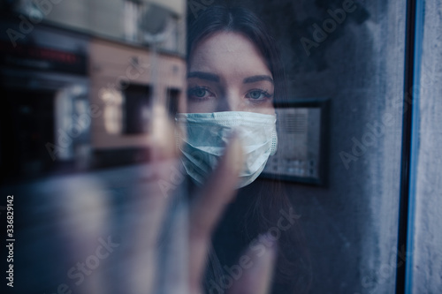 Coronavirus. Sick woman of covid-19 looking through the window and wearing mask protection. Patient isolated to prevent infection during Quarantine. The woman wears a mask because of air pollution.